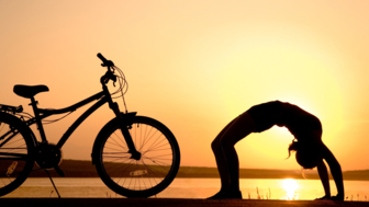 Eine Mountainbikerin in einer Yogaposition in einer Brückenhaltung mit dem Mountainbike nebenan bei Sonnenuntergang an einem See.