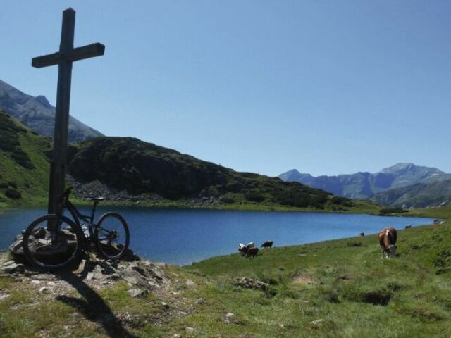 Ein Mountainbike an einen Gipfelkreuz angelehnt mit Blick auf einen Alpensee