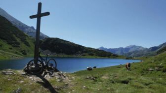 Ein Moutainbike steht am Gipfelkreuz mit einem Alpensee im Hintergrund.