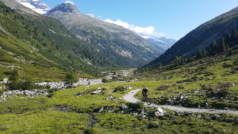 Ein Mountainbiker unterwegs auf der Transalp vom Tegernsee zum Molvenosee mit der Bergen im Hintergrund entlang eines Bachlaufes fahrend.