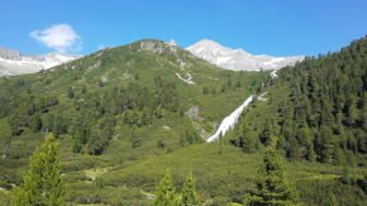 Ein Wasserfall schlängelt sich zwischen dem Bergwald in die Tiefe mit der umliegenden Bergwelt im Hintergrund.